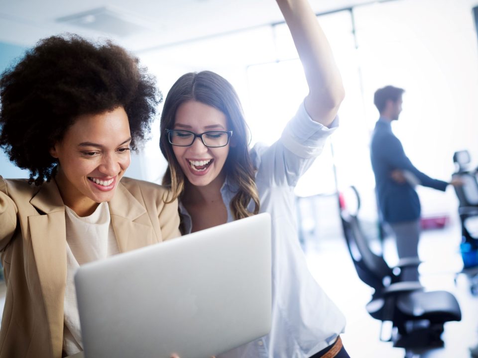 Implementing employee engagement ideas and programs can help boost morale, as represented by this image of two women celebrating success while looking at a laptop.